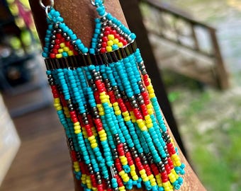 May include: Turquoise, red, yellow, and silver beaded earrings with a fringe design. The earrings have a triangular top with a silver metal bar and a beaded fringe. The earrings are hanging on a brown surface.