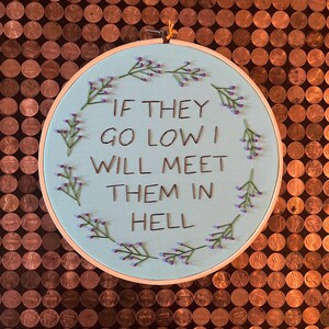 May include: A light blue embroidery hoop with the text "If they go low I will meet them in hell" surrounded by purple flowers.