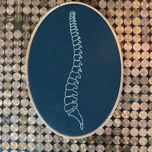 Pode incluir: Um bastidor de bordado oval com um desenho de coluna vertebral costurado em branco sobre tecido azul escuro. O bastidor tem uma moldura em tom de madeira natural. O fundo é um mosaico de moedas.