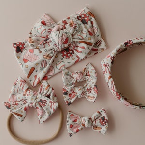 May include: A set of five hair accessories featuring a baseball theme. The set includes a headband, two large hair bows, a smaller hair bow, and a thin elastic headband. All accessories are white with red and brown baseball-themed illustrations.