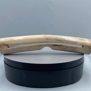 Op de afbeelding: Een gebogen, gebroken wit benen object met twee parallelle sleuven en een centrale inkeping. Het bot heeft natuurlijke kleurvariaties, met donkerdere gebieden aan de uiteinden. Het object wordt gepresenteerd op een zwart-grijs draaiend platform.