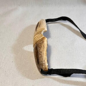May include: A carved, light brown pendant with a curved design and a black cord. The pendant has a textured surface with carved lines. The black cord is attached to the pendant and appears to be made of a woven material.