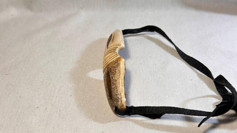 May include: A carved, light brown pendant with a curved design and a black cord. The pendant has a textured surface with carved lines. The black cord is attached to the pendant and appears to be made of a woven material.