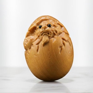 May include: A detailed wooden carving of a mythical beast hatching from an egg. The sculpture is light brown with black eyes. The carving features a dragon-like face and textured eggshell.