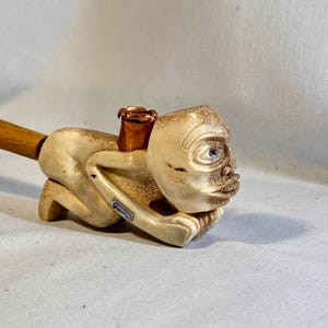 Pode incluir: Um cachimbo único com uma figura esculpida com rosto humano e um cabo de madeira. A figura é creme com traços faciais detalhados e uma tigela cor de cobre. O cabo é de cor castanho claro.