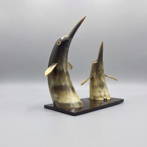 May include: A decorative sculpture featuring two stylized swordfish figures crafted from polished horn material. The figures exhibit natural brown, cream, and black marbled patterns, mounted on a rectangular dark brown wooden base stand.