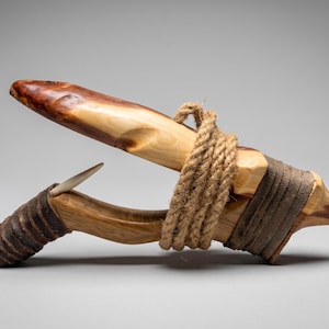 May include: A handcrafted wooden fishhook featuring a polished light and dark brown wood body, secured with thick tan jute rope wrapping. A sharp, light-coloured bone or antler point is embedded into the lower section of the curved wooden frame.