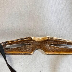 May include: A handcrafted, brown eye mask with a natural wood grain appearance. The mask features two eye openings and is connected to black fabric straps for securing around the head. The mask is designed for sleep or relaxation.