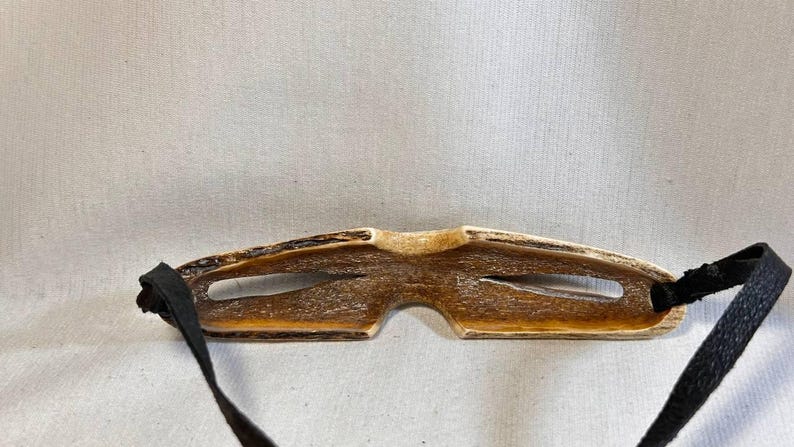 May include: A handcrafted, brown eye mask with a natural wood grain appearance. The mask features two eye openings and is connected to black fabric straps for securing around the head. The mask is designed for sleep or relaxation.