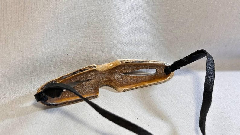 May include: A brown and tan antler eyeglass holder with a black strap. The holder has a unique shape with a cutout and is designed to hold eyeglasses. The black strap is attached to the holder.