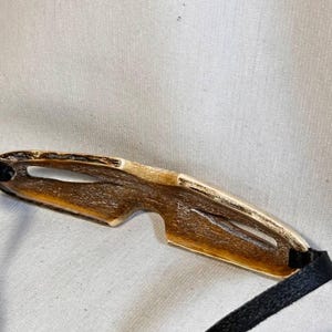May include: A pair of handcrafted, unique sunglasses with a natural, organic aesthetic. The frame is a warm, light brown with a golden trim, and features a distinctive, open design. Black fabric straps are attached to the frame.