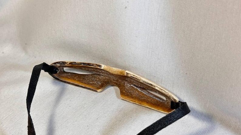 May include: A pair of handcrafted, unique sunglasses with a natural, organic aesthetic. The frame is a warm, light brown with a golden trim, and features a distinctive, open design. Black fabric straps are attached to the frame.