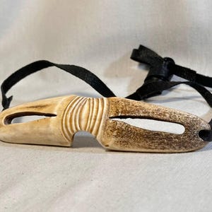 May include: A carved, off-white eye mask with a dark brown, aged appearance. The mask has a curved shape with two eye openings and is attached to a black leather strap. The mask has a textured, ridged design.