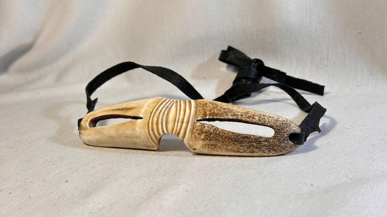 May include: A carved, off-white eye mask with a dark brown, aged appearance. The mask has a curved shape with two eye openings and is attached to a black leather strap. The mask has a textured, ridged design.