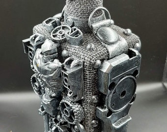 Può includere: Bottiglia decorativa argentata con design steampunk. Il collo è avvolto in tessuto strutturato. Il corpo è decorato con ingranaggi, parti meccaniche e volti simili a robot, creando un'estetica industriale unica.