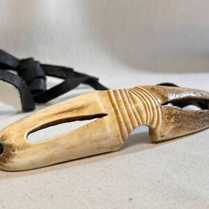 May include: A carved pendant with a natural bone appearance, featuring a central oval opening and a textured, ridged design on one end. It is attached to a black leather cord.