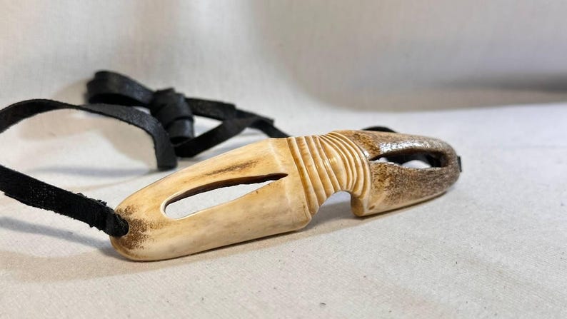 May include: A carved pendant with a natural bone appearance, featuring a central oval opening and a textured, ridged design on one end. It is attached to a black leather cord.