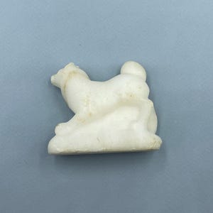 Könnte beinhalten: Kleine, cremefarbene Hundefigur in sitzender Position. Der Hund ist einfach modelliert und sitzt auf einem kleinen Sockel. Die Figur hat ein leicht gealtertes Aussehen mit subtilen Farbvariationen. Die Basis ist etwa 3 cm lang.