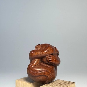 May include: A polished dark brown wooden sculpture depicting a figure in a curled, fetal position with arms wrapped around the head and knees. The wood grain is visible across the smooth, rounded surface of the carved form, showing intricate details.
