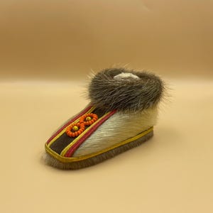 Könnte beinhalten: Ein einzelner Fellschuh mit braunem Fellbesatz. Der Schuh hat einen cremefarbenen Fellkörper, der mit roten und gelben Stickereidetails versehen ist. Die Sohle ist mit gelben Nähten umrandet.