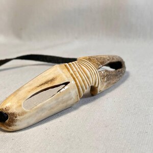 May include: A handcrafted, off-white antler pendant with a carved design and a black cord. The pendant has a unique shape with a hole at one end and a carved section in the middle. The cord is attached to the pendant.
