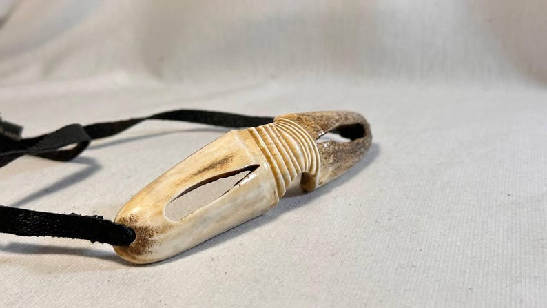 May include: A handcrafted, off-white antler pendant with a carved design and a black cord. The pendant has a unique shape with a hole at one end and a carved section in the middle. The cord is attached to the pendant.