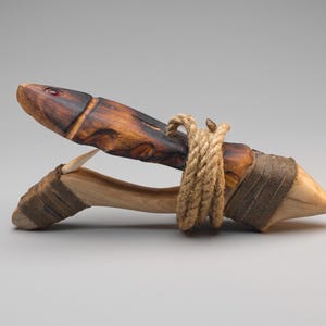 Può includere: Un'esca in legno intagliato a forma di pesce con la parte superiore tinta di marrone scuro e un piccolo occhio rosso, fissata a una base in legno più chiaro con una corda spessa color cuoio e un amo curvo e affilato che sporge dal centro.