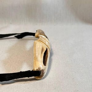 May include: A handcrafted bone pendant with a black leather cord. The pendant is rectangular with a carved design and a central opening. The cord is approximately 12 inches long.