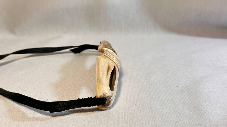 May include: A handcrafted bone pendant with a black leather cord. The pendant is rectangular with a carved design and a central opening. The cord is approximately 12 inches long.