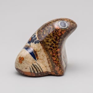 Può includere: Scultura in ceramica di una testa di pesce. La scultura presenta un corpo marrone e beige maculato con accenti floreali blu e arancioni. Il pesce ha un singolo occhio rotondo blu e bianco.
