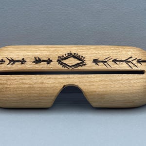 May include: A light-colored wooden glasses case with a decorative tribal design. The case has a curved shape with a cutout for the nose bridge and a slot for the glasses. The design includes arrows and a diamond shape.