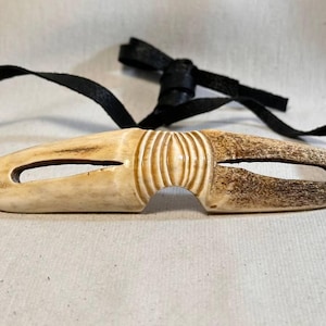 May include: A carved, bone-colored mask with a black strap. The mask has a central, ridged design and two eye openings. The right side of the mask is a darker brown color. The black strap is tied at the top.
