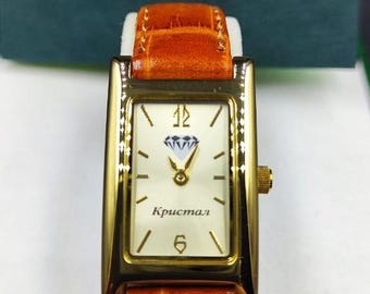 May include: A rectangular gold-toned wristwatch with a white face, featuring black hour markers and hands. The watch has a diamond-shaped design at the 12 o'clock position and the word "Кристал" on the face. It has a brown leather band.