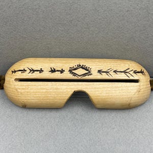 May include: A light-colored wooden slit drum instrument with a smooth, rounded rectangular shape. It features a central horizontal slit, a small arched cutout at the bottom, and dark, hand-burned arrow and diamond patterns. Brown leather straps are attached.