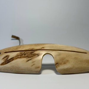 May include: A curved, light-colored wooden eye mask featuring a natural wood grain pattern with darker brown swirls. The mask has a central nose cutout, a horizontal slit across the front, and black leather straps attached at both sides.
