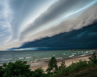 May include: A dark, ominous storm cloud hangs low over a calm, blue ocean and sandy beach. The sky is a dramatic mix of dark gray and light blue, with the cloud casting a shadow over the water.