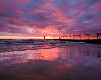 May include: A long pier extends into the water at sunset, with a lighthouse in the distance. The sky is a vibrant mix of pink, purple, and orange, reflecting in the calm water.