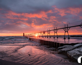 May include: A long pier extends into the water at sunset, with a lighthouse in the distance. The sky is a vibrant mix of pink, orange, and purple, with clouds reflecting the colors. The water is choppy and the pier is silhouetted against the sky.