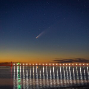 May include: A red and white lighthouse stands on a pier with lights illuminating the water. A comet streaks across the night sky.