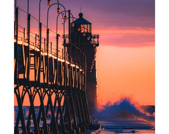 May include: A silhouette of a lighthouse on a pier with a dramatic sunset in the background. The pier is made of wood and has a metal railing. The waves are crashing against the pier, creating a spray of water.