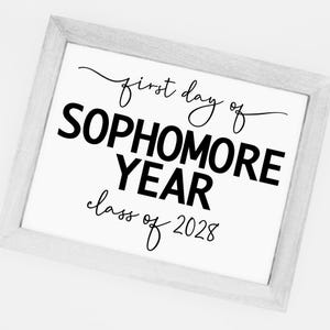 May include: A framed white sign with black text that reads "first day of SOPHOMORE YEAR class of 2028". The frame is a light gray color.