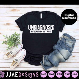 Op de afbeelding: Een zwart T-shirt met witte tekst die "Undiagnosed but something ain't right" luidt. Het shirt ligt op een houten oppervlak met een paar witte sneakers en een blauwe jeans.