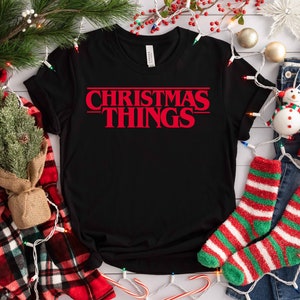 Puede incluir: Camiseta negra con el texto rojo "CHRISTMAS THINGS" en una fuente retro. La camiseta está rodeada de decoraciones navideñas, incluyendo una manta a cuadros, calcetines y una taza que dice "Merry Christmas".