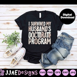 May include: A black t-shirt with the text "I survived my husband's doctorate program" in a light brown colour. The shirt is laid out on a wooden surface with a pair of white trainers, a pair of blue jeans, and a potted plant.