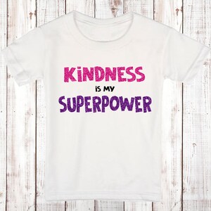 Puede incluir: Camiseta blanca con el texto "KINDNESS is my SUPERPOWER" en letras de purpurina rosa y morada.