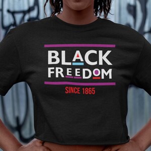 Könnte beinhalten: Schwarzes Crop-Top mit dem Text "BLACK FREEDOM SINCE 1865" in weißen Buchstaben mit bunten Akzenten auf schwarzem Hintergrund.