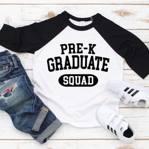 May include: White and black baseball tee shirt with the text "PRE-K GRADUATE SQUAD" in black. The shirt is on a white wooden surface with a pair of blue jeans and white sneakers.
