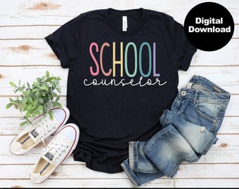 以下が含まれることがあります： レインボーカラーで「SCHOOL counselor」と書かれた黒いTシャツ。遊び心のあるフォントで書かれています。Tシャツは、白いスニーカーと青いジーンズと一緒に木製の表面に置かれています。