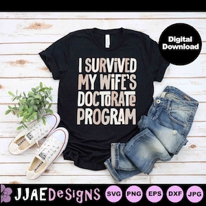 May include: A black t-shirt with the text "I survived my wife's doctorate program" in a light brown color. The shirt is laid out on a wooden surface with a pair of white sneakers, blue jeans, and a green plant.