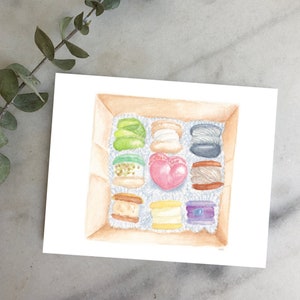 May include: A watercolor painting of a box of assorted macarons. The box contains nine macarons, including a heart-shaped macaron, a green macaron with white sprinkles, a black macaron, and a white macaron with a pink heart.